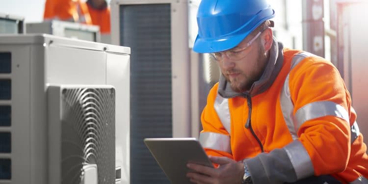 An air conditioning engineer is finishing the installation of several units on a rooftop. Two colleagues can be seen also installing units in the background. They are wearing hi vis jackets, hard hats and safety goggles.