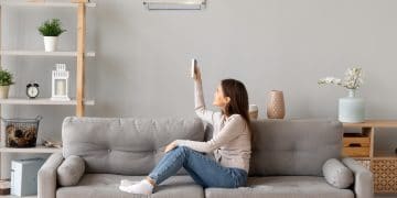 Young smiling woman in casual wear sitting on couch in living room resting holding remote controller switch on air conditioner make regulate comfort temperature at home enjoy fresh or warm air concept.