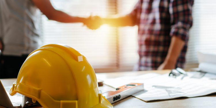 yellow safety helmet on workplace desk with construction worker team hands shaking greeting start up plan new project contract in office center at construction site, partnership and contractor concept
