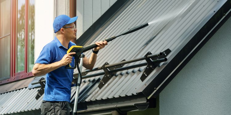 man standing on ladder and cleaning house metal roof with high pressure washer