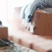 Male builder hand in gloves holding clay brick on background of brickwork wall. Repair and redevelopment apartments concept.