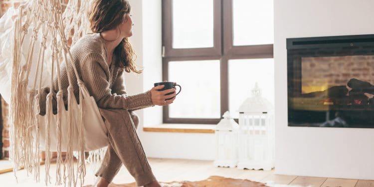 Beautiful young woman, which is wearing cashmere nightwear clothes, drinking tea or coffee while relaxing on rocking chair in scandinavian cabin near electric heater fireplace one lazy weekend morning