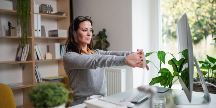 An attractive businesswoman sitting indoors in office, stretching.