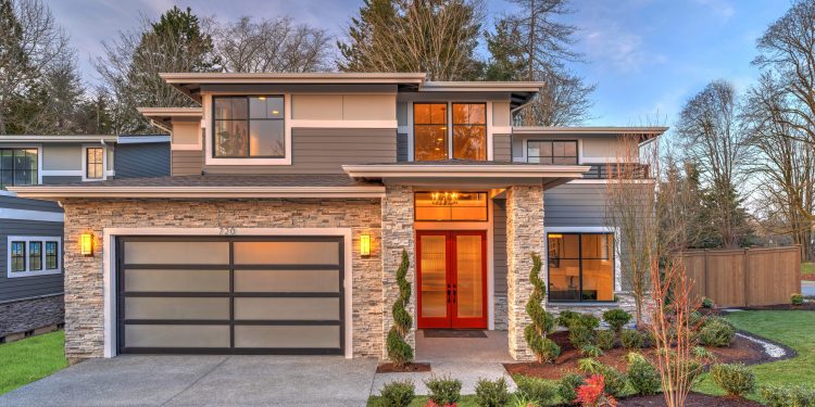 Modern contemporary house exterior with luxury details, landscaping, stone, wood, glass, lots of large windows with evening lights and red front door.