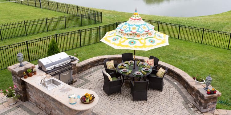 Outdoor living space on a brick patio overlooking a tranquil lake and fenced green lawn with a table under a sunshade or umbrella laid ready for dinner, high angle view