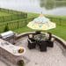 Outdoor living space on a brick patio overlooking a tranquil lake and fenced green lawn with a table under a sunshade or umbrella laid ready for dinner, high angle view