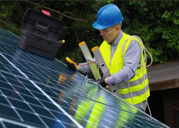 A person wearing a hard hat and safety vest working on a solar panel Description automatically generated with low confidence